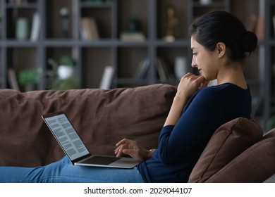 Happy millennial Indian woman sit relax on couch use computer study online from home. Smiling young mixed race female rest on sofa work distant on laptop prepare research on web.