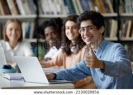 Happy millennial Jewish male student in eyeglasses showing thumbs up gesture, using computer, working on online project or preparing for exams in college library with smiling multiracial friends.