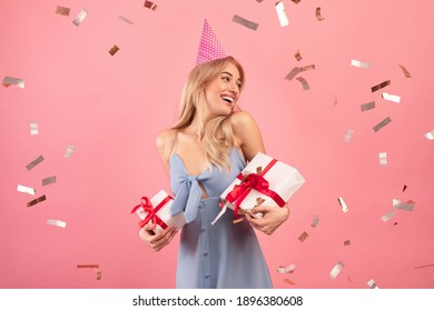 Happy millennial lady holding wrapped gift boxes, wearing birthday hat on pink studio background with falling confetti. Lovely blonde having B-day party, clubbing with presents. Festive concept