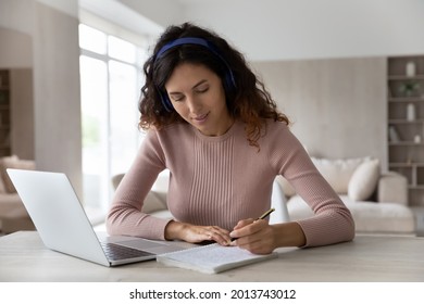 Happy Millennial Latino Female Student In Earphones Sit At Desk At Home Write Take Notes Study Online On Laptop. Young Hispanic Woman In Headphones Handwrite Work Distant On Web On Computer.