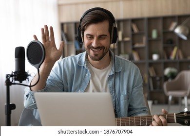 Happy Millennial Male Artist In Headphones And Guitar Have Online Video Music Lesson On Computer. Smiling Young Man Singer Tutor Or Coach Talk Greet On Webcam Digital Virtual Training At Home Studio.