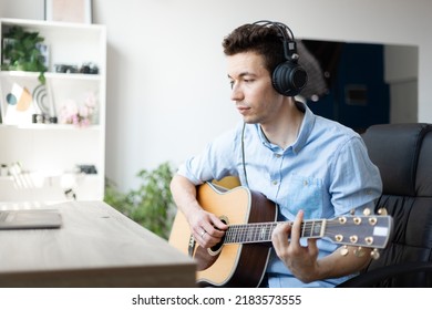 Happy Millennial Male Artist In Headphones And Guitar Have Online Video Music Lesson On Computer. Practicing Guitar In Living Room. 