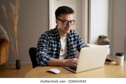 A happy millennial man with glasses, a webinar or a training course or a computer, studying online from home. Programmer guy with glasses is sitting at his desk, working on a laptop, listening to musi - Shutterstock ID 2246514543