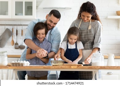 Happy Millennial Married Couple Teaching Small Kids Making Homemade Pastry. Smiling Adorable Little Children Enjoying Preparing Dough With Excited Parents In Modern Kitchen, Family Holiday Activity.