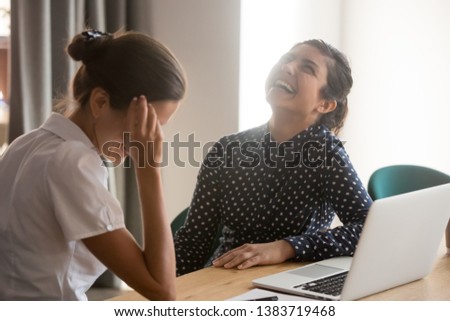 Happy millennial multiethnic women have fun sitting at office desk talking, smiling diverse female colleagues or employees laugh chatting at workplace, discuss ideas or gossip at work break