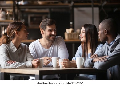 Happy Millennial Multiracial Friends Sit In Coffeeshop Enjoying Hot Coffee In Mugs, Smiling Young Diverse People Have Fun Meeting Hanging Out In Cafe Talking Chatting Drinking Tea Beverage