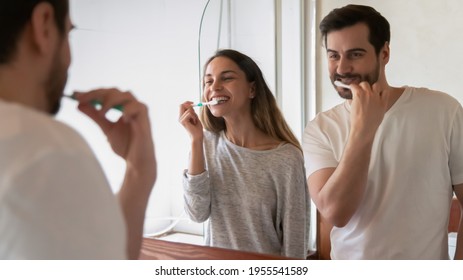 Happy millennial multiracial newlyweds couple look in mirror in new home bathroom brush teeth together. Smiling young man and woman enjoy early morning washing routine in bath. Rental concept.