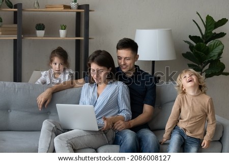 Happy millennial parents relaxing on couch, using online app on laptop, active naughty sibling kids walking around, having fun, laughing. Family couple and children using computer, enjoying leisure