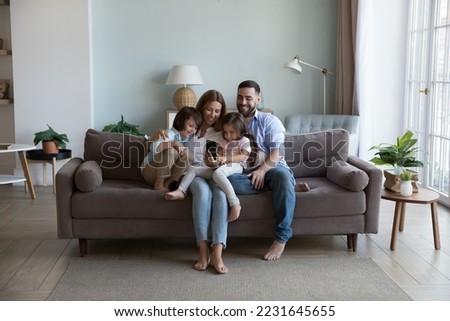 Happy millennial parents and two kids using mobile phone, resting on home couch together, holding smartphone, taking selfie, relaxing in living room interior. Family communication concept