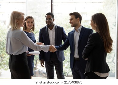 Happy millennial team leader handshaking and greeting warmly senior female executive with personal achievement, promotion, success at informal meeting at lunch break in presence of diverse colleagues - Powered by Shutterstock