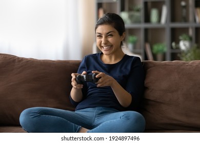 Happy Millennial Teen Indian Female Sit Relax On Sofa At Home Have Fun Play Online Video Game. Overjoyed Young Mixed Race Woman Gamer Involved In Virtual Reality With Videogames. Hobby Concept.