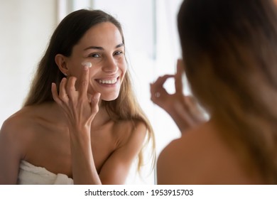 Happy millennial teenage Latin woman look in mirror in bathroom apply nourishing moisturizing cream for health glowing face skin. Smiling young Hispanic female do beauty facial procedures in bath. - Powered by Shutterstock