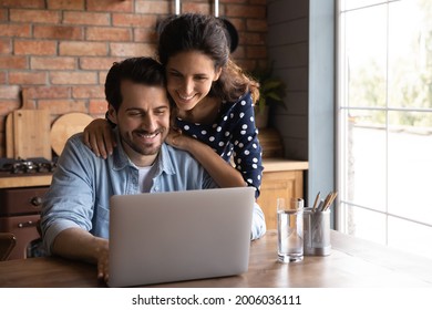 Happy Millennial Wife Giving Support To Husband Working At Laptop From Home, Touching Shoulders. Married Couple Surfing Internet On Computer Together, Paying Bills Online, Reviewing Mortgage Terms