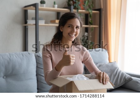 Happy millennial woman holding carton box on lap, resting on comfortable couch, showing thumbs up gesture. Smiling young female client looking at camera, recommending purchases in internet store.