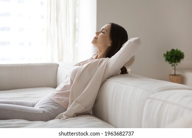 Happy millennial woman relaxing on sofa, breathing cool air from conditioner, enjoying comfortable climate in ventilated living room, meditating with closed eyes. Comfort at home, conditioning concept