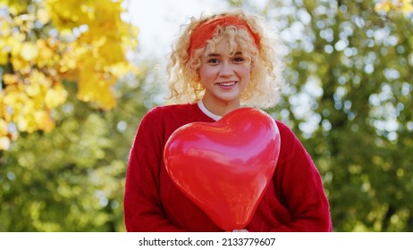 Happy Millennial Young Girl With Blond Curly Hair In Red Holding Heart Baloon - Medium Shot. High Quality Photo