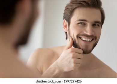 Happy Millennial Young Handsome Man Looking In Mirror In Bathroom With Toothy Smile, Scanning Facial Skin And Stylish Stubble. Guy Satisfied With Male Beauty Care Product. Skincare, Home Spa Concept