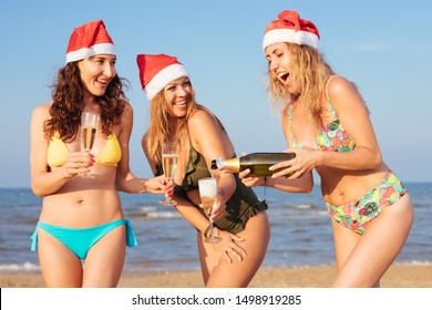 Happy millennials girls celebrating Christmas on the beach drinking champagne, wearing Santa's red hats - young smiling woman pouring prosecco wine to her girlfriends - xmas beach party concept