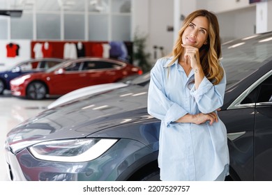 Happy minded dreamful thoughtful woman customer female buyer client in blue shirt choose auto want to buy new automobile in car showroom vehicle salon dealership store motor show indoor. Sales concept