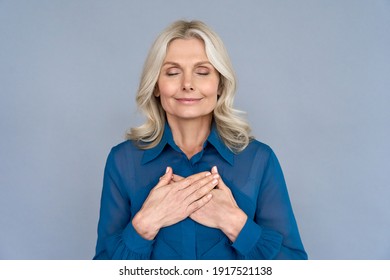 Happy Mindful Thankful Middle Aged Old Woman Holding Hands On Chest Meditating With Eyes Closed Isolated On Grey Background Feeling No Stress, Gratitude, Mental Health Balance, Peace Of Mind Concept.