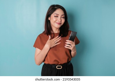 Happy mindful thankful young woman holding phone and hand on chest smiling isolated on blue background feeling no stress, gratitude, mental health balance, peace of mind concept. - Shutterstock ID 2193343035