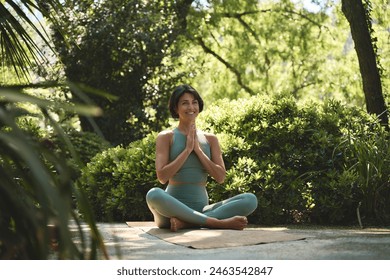 Happy mindful young Hispanic woman meditating doing yoga breathing exercises feeling peace of mind, mental balance sitting in lotus pose hands in namaste in green nature park outdoors. - Powered by Shutterstock