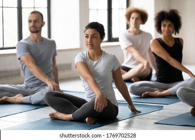 Happy Mindful Young Mixed Race People Doing Simple Seated Twist Exercise, Multiethnic Diverse Beginners Practicing Parivrtta Sukhasana Barefoot On Yoga Mat At Group Class In Modern Interior.