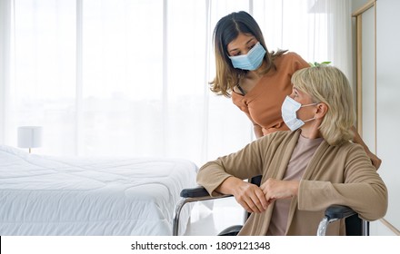 Happy mix skin family living together, asian daughter takes care of her adopted caucasian mother sitting on a wheelchair. Both wearing face mask.