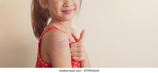 Happy Mixed Asian Girl  Giving Thumb Up And Showing Her Arm With Bandage After Got Vaccinated Covid Delta Or  Inoculation, Child Immunization