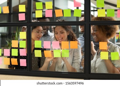 Happy Mixed Race Colleagues Watching Smiling Team Leader Marking Task As Done On Colorful Sticky Notes Of Kanban Agile Glass Window Board At Office. Diverse Teammates Satisfied With Working Process.