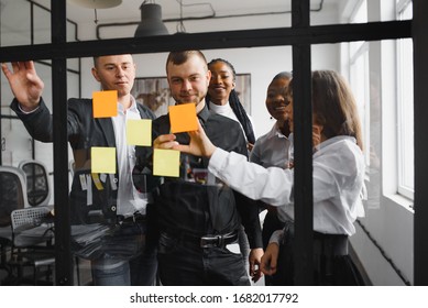 Happy Mixed Race Colleagues Watching Smiling Team Leader Marking Task As Done On Colorful Sticky Notes Of Kanban Agile Glass Window Board At Office. Diverse Teammates Satisfied With Working Process.