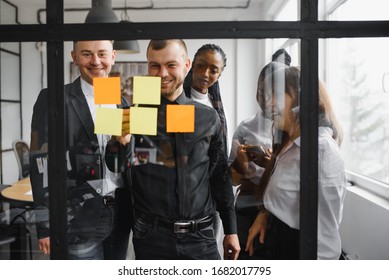 Happy Mixed Race Colleagues Watching Smiling Team Leader Marking Task As Done On Colorful Sticky Notes Of Kanban Agile Glass Window Board At Office. Diverse Teammates Satisfied With Working Process.