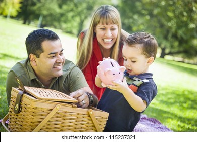 Happy Mixed Race Couple Give Their Son a Piggy Bank at a Picnic in the Park.