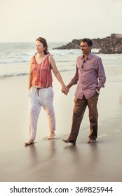 Happy Mixed Race Couple Holding By Hand And Walking On The Beach At Sunset. 