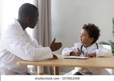 Happy Mixed Race Cute Little Preschool Boy Sitting At Table, Wearing Doctor Uniform, Pretending To Be General Practitioner, Playing With Friendly African American Male Pediatrician At Hospital.