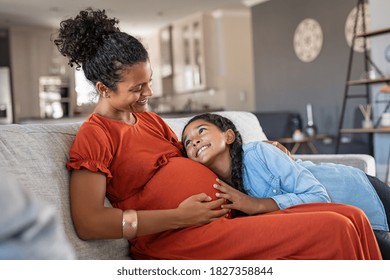 Happy mixed race daughter hugging belly of her expecting mother at home. African girl listening to baby movements while embracing pregnant woman. Pregnant black mom and future sister relaxing on sofa.