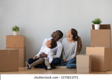 Happy Mixed Race Family Of African American Father, Mother And Cute Little Curious Son Sitting On Floor Between Big Cardboard Boxes, Inspecting New Apartment, Discussing First House Renovation Ideas.