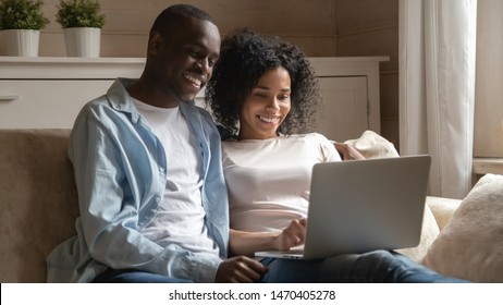 Happy mixed race family couple relaxing on comfortable couch at home, using laptop, enjoying leisure weekend time together, watching movies, shopping in popular internet store, chatting with friends. - Powered by Shutterstock