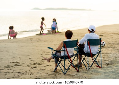 Happy Mixed Race Family Holiday Vacation. Grandparents Sitting On Outdoor Chair Looking At Cute Mixed Race Grandchild Girl On The Beach In Summer Day. Family Relax And Having Fun In Summer Weekend