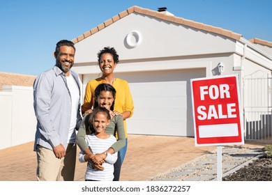 Happy Mixed Race Family Standing Outside Home With Sale Signboard. Couple With Two Daughters Standing By For Sale Sign Outside Their New House While Looking At Camera, Put The House Up For Sale.