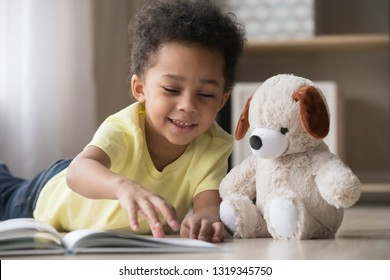 Happy Mixed Race Little Boy Enjoying Playing Alone Reading Book To Fluffy Toy Lying On Warm Floor, Cute Smiling African American Kid Having Fun At Home, Creative Child Activity, Underfloor Heating