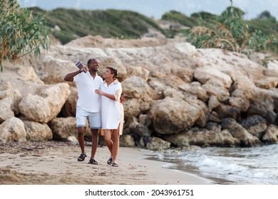 Happy Mixed Race, Middle Age Couple Embracing While Walking On The Beach Holding A Guitar. Man Holding Guitar With Woman.