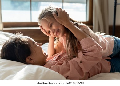 Happy mixed race mother lying on bed, stroking head of little adopted kid daughter. Smiling young woman looking at eyes of adorable small child girl, enjoying sweet tender moment in bedroom at home.