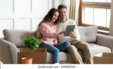 Happy Mixed Race Woman Sitting On Couch With Pleasant Caucasian Man, Using Digital Tablet, Ordering Items Furniture Online After Moving In New House. Smiling Family Couple Shopping In Internet.