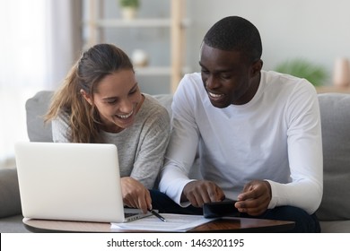 Happy mixed race young couple calculate domestic bills mortgage rate bank loan payment at home, smiling interracial family planning budget manage household expenses paperwork use laptop calculator