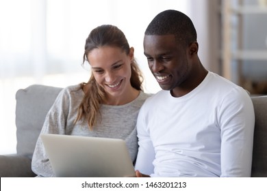 Happy Mixed Race Young Couple Using Laptop Looking At Screen Together Watching Video Movie Sit On Sofa, Smiling Interracial Husband And Wife Make Online Computer Call Do Internet Shopping At Home