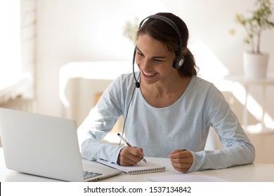 Happy mixed race young woman wearing headset, looking at laptop screen, watching educational lecture, enjoying interesting webinar, writing notes. Smiling employee holding video call with clients.