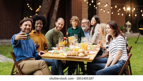 Happy Mixed-races Family Sitting In Back Yard At Table And Having Dinner. Multi Ethnic Cheerful People Taking Selfie Photo With Smartphone Outdoor. Mobile Phone Picture. 