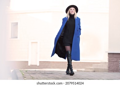 Happy model with beautiful smile walking street in motion. Blonde woman wearing fedora, blue autumn or spring coat, black knitted sweater, shorts, high leather boots. Light background