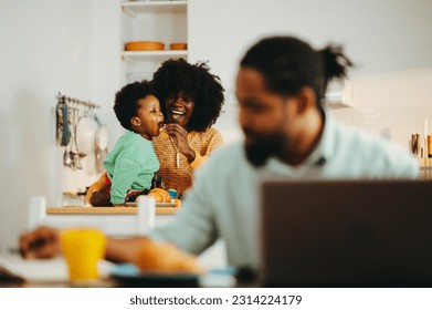 A happy modern african american family at home. The mother is feeding her son in the kitchen while the busy father is finishing his remote work on a laptop and writing down in an agenda. - Powered by Shutterstock